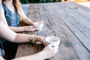 2 women sitting at a wooden table holding cups of coffee