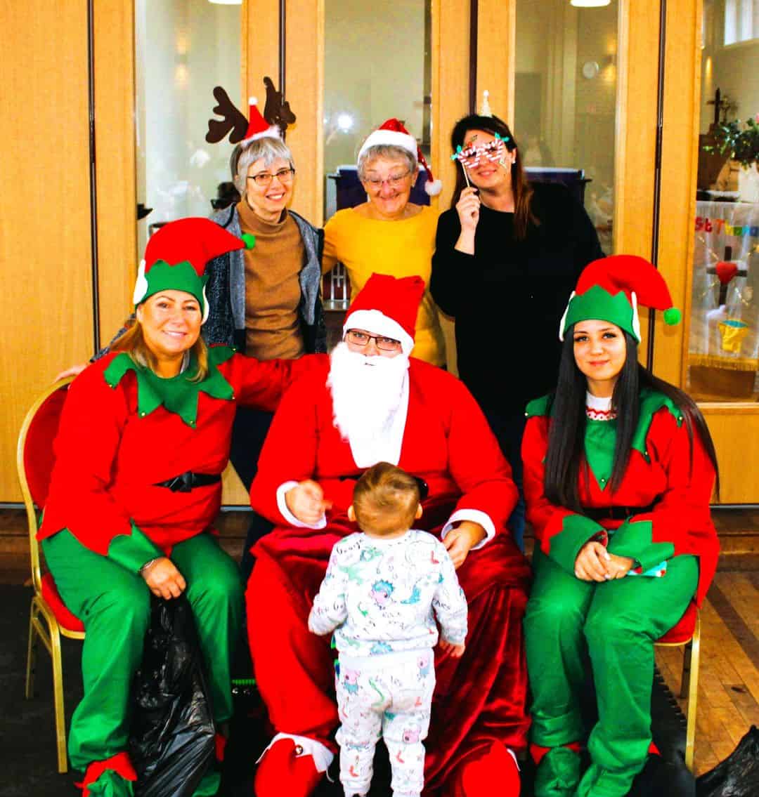 Santa and 2 elves siting giving a present to a small child